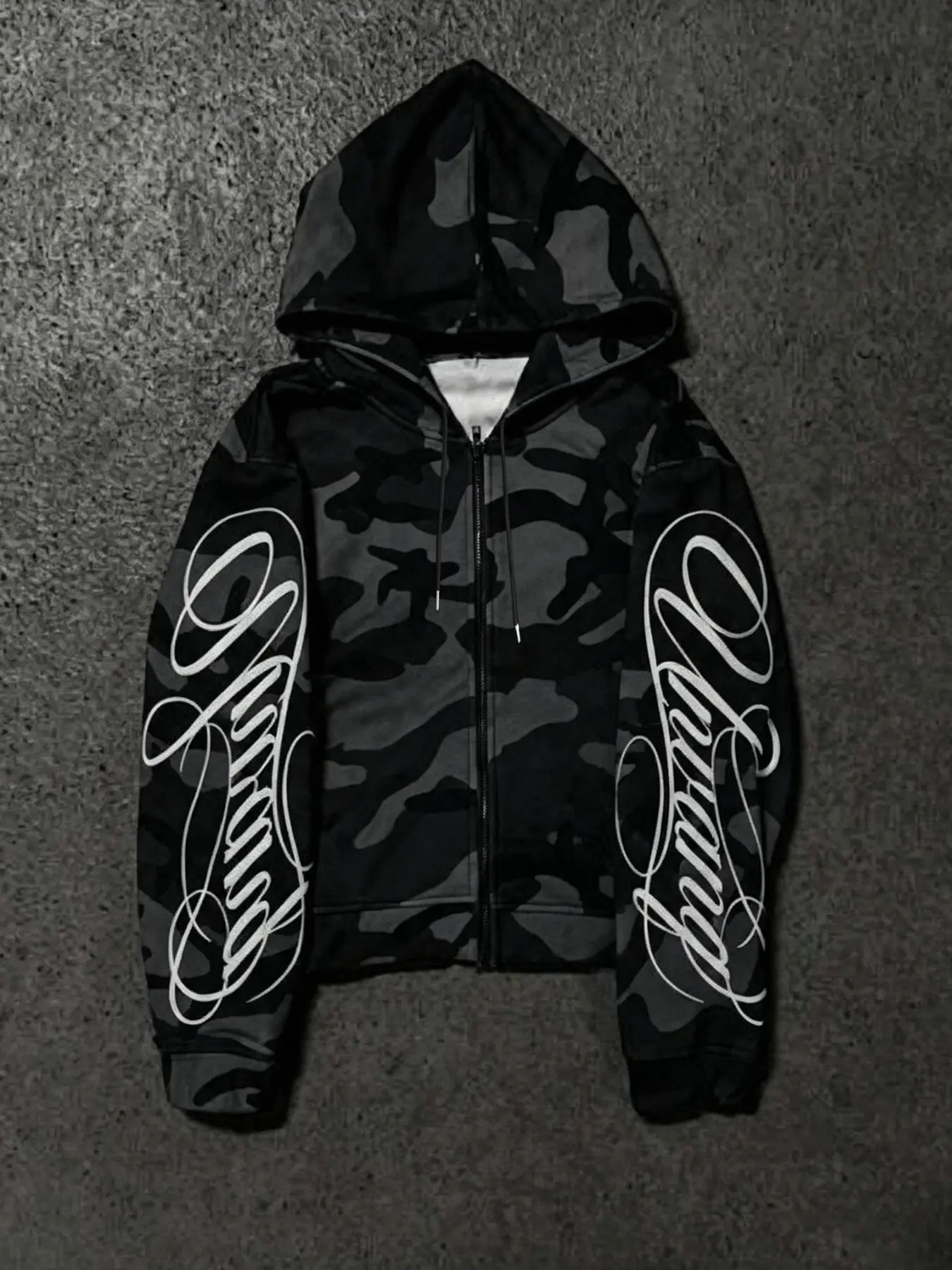 Black-gray camo zip-up hooded jacket with white cursive prints on both sleeves. Oversized loose fit, trendy street style, soft fabric, versatile and cool for daily casual outfits.