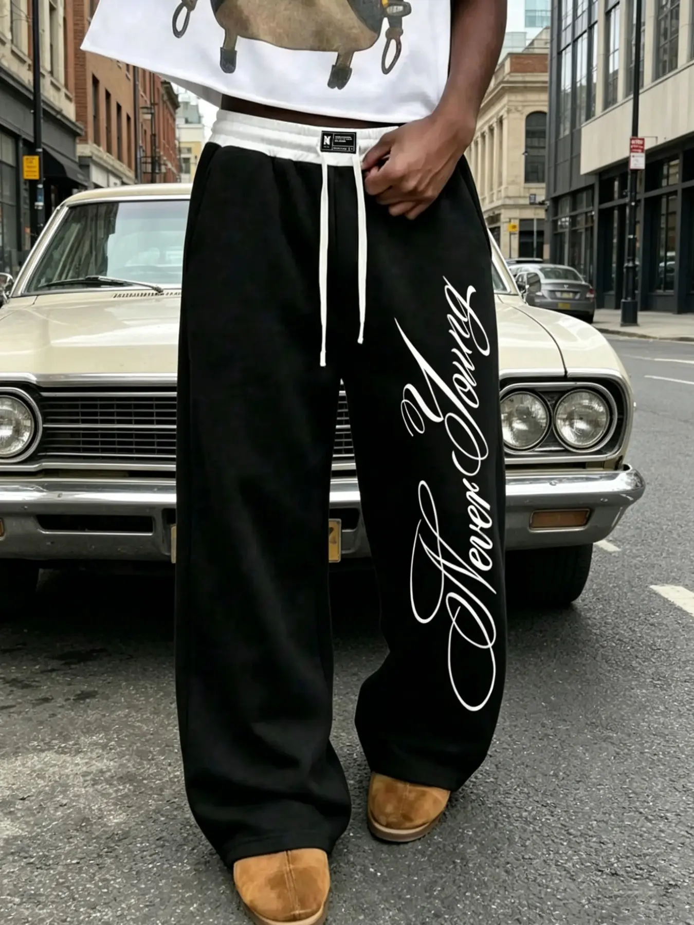 Men's Solid Black Wide-Leg Sweatpants, Loose Hip-Hop Sports Trousers. Features contrasting white ribbed waistband paired with two white drawcords and woven labels. Vertical cursive handwritten print of the English phrase "Never Young" runs down the side o